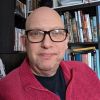 Headshot of Richard Hill FAHACS MA student sat in front of a bookshelf. Richard wears a black t shirt and red fleece.
