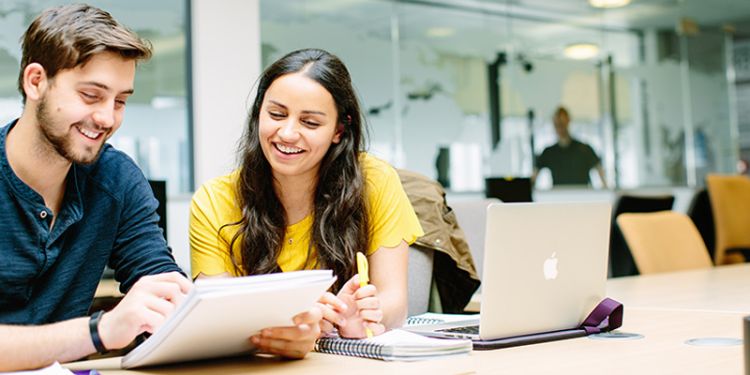 Two people reviewing a piece of work on a paper while sat at a desk with a laptop and notebook.