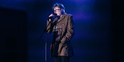 Person wearing glasses on a dark stage stalking into a microphone