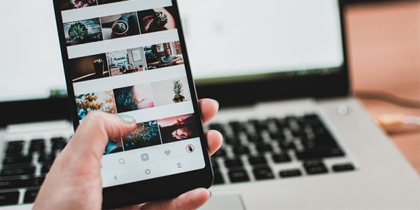 An image of someone using a phone to scroll through photos, with a laptop turned on in the background.
