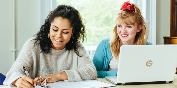 Two students smiling and working with laptop and notebook/pen.