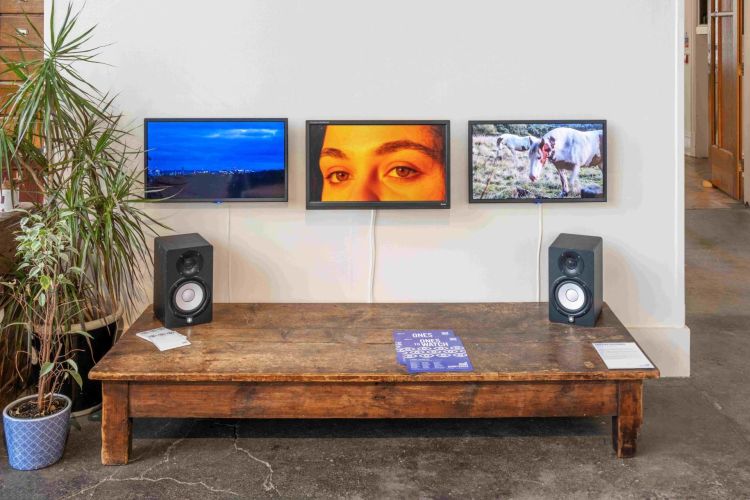 Audio visual installation by artist Angelika Kaccouris consisting of three video screens are side by side on a wall. Underneath is a table  holding two speakers.